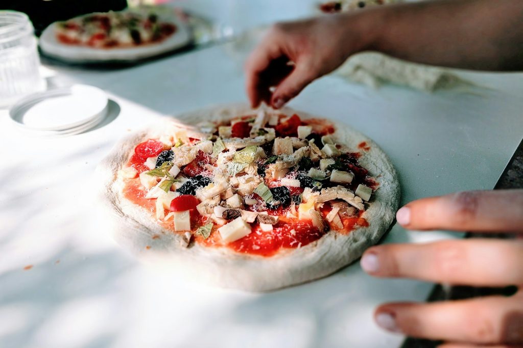 Fresh pizza being prepared on a floured counter with tomato sauce, cheese, and toppings in natural light