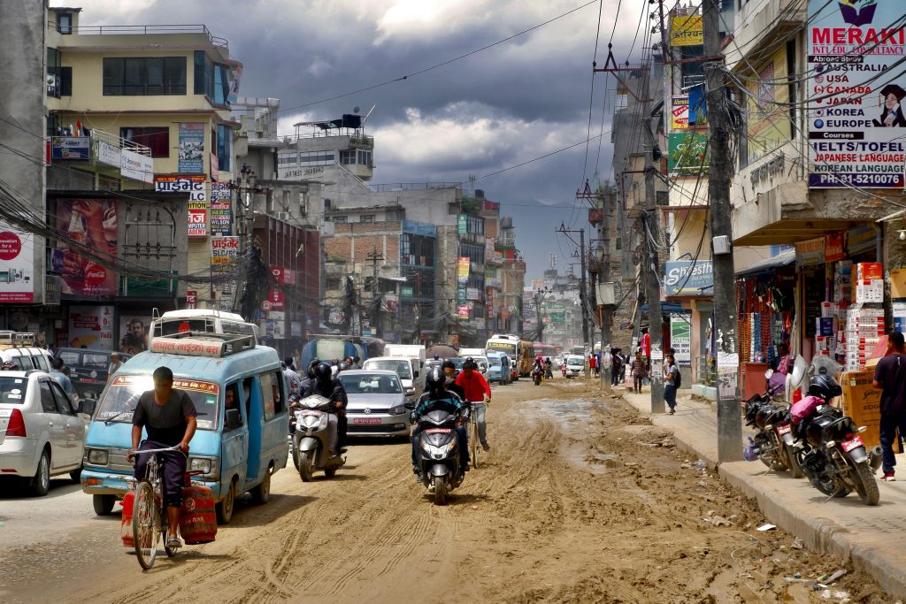 Busy street scene in Kathmandu, Nepal, with traffic, motorbikes, shops, and tangled overhead power lines.
