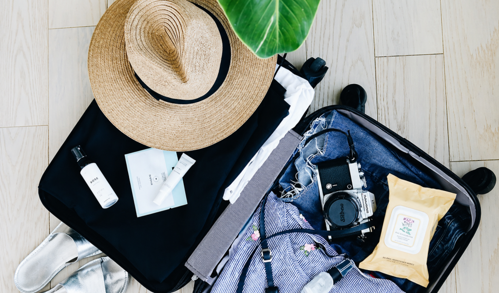 Open suitcase with clothes, camera and travel accessories next to a straw hat on a light floor